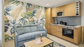 Un espacio habitable compacto con kitchenette, un sofá gris, una mesa de centro y un papel tapiz con estampado de hojas tropicales.