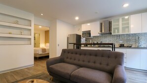 Una sala de estar moderna con un sofá gris, una mesa de centro de madera y una cocina con armarios blancos y un microondas.