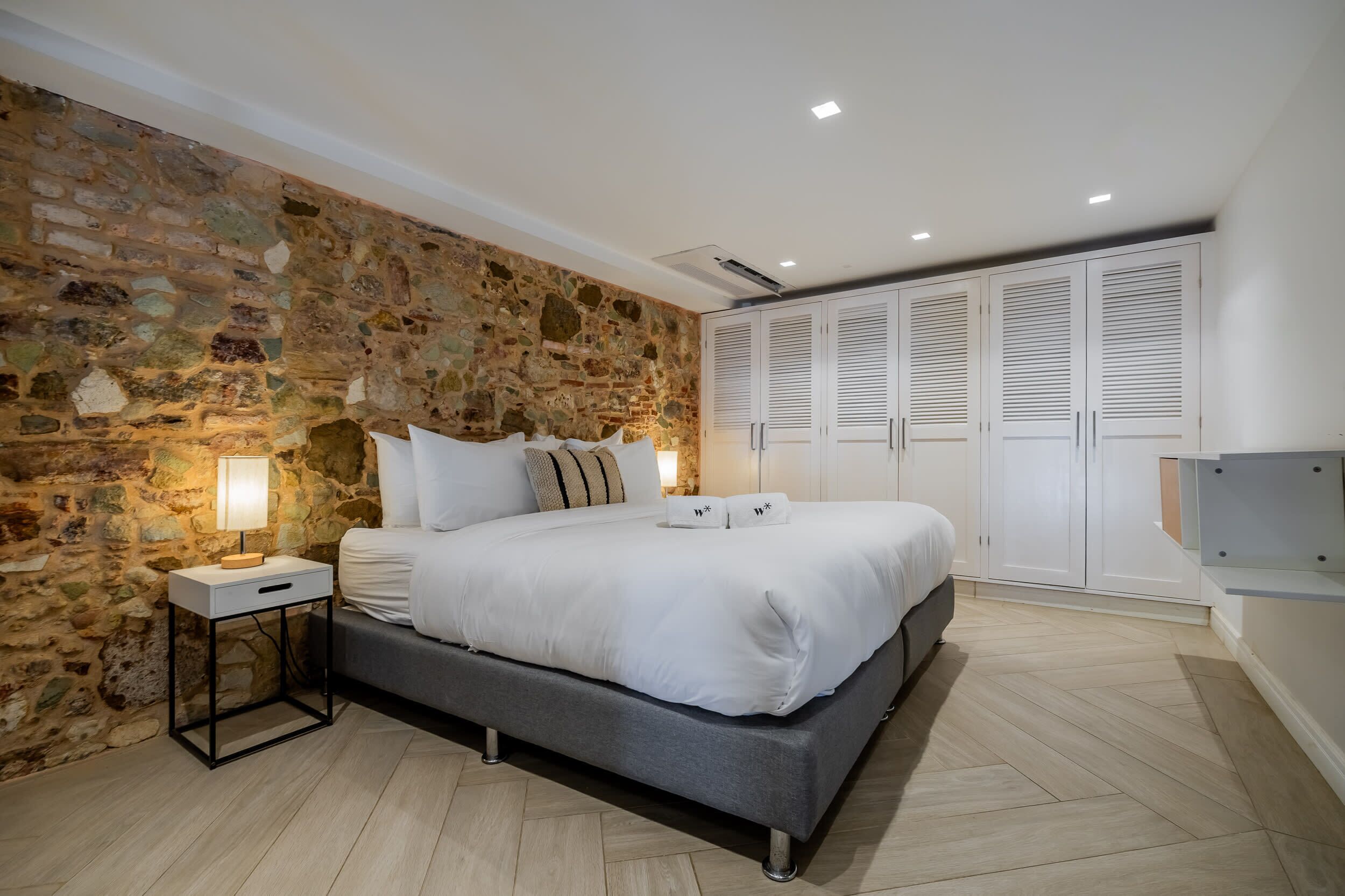 A bedroom with a stone wall, a bed with white bedding, a bedside table with a lamp, and a wardrobe with white doors.