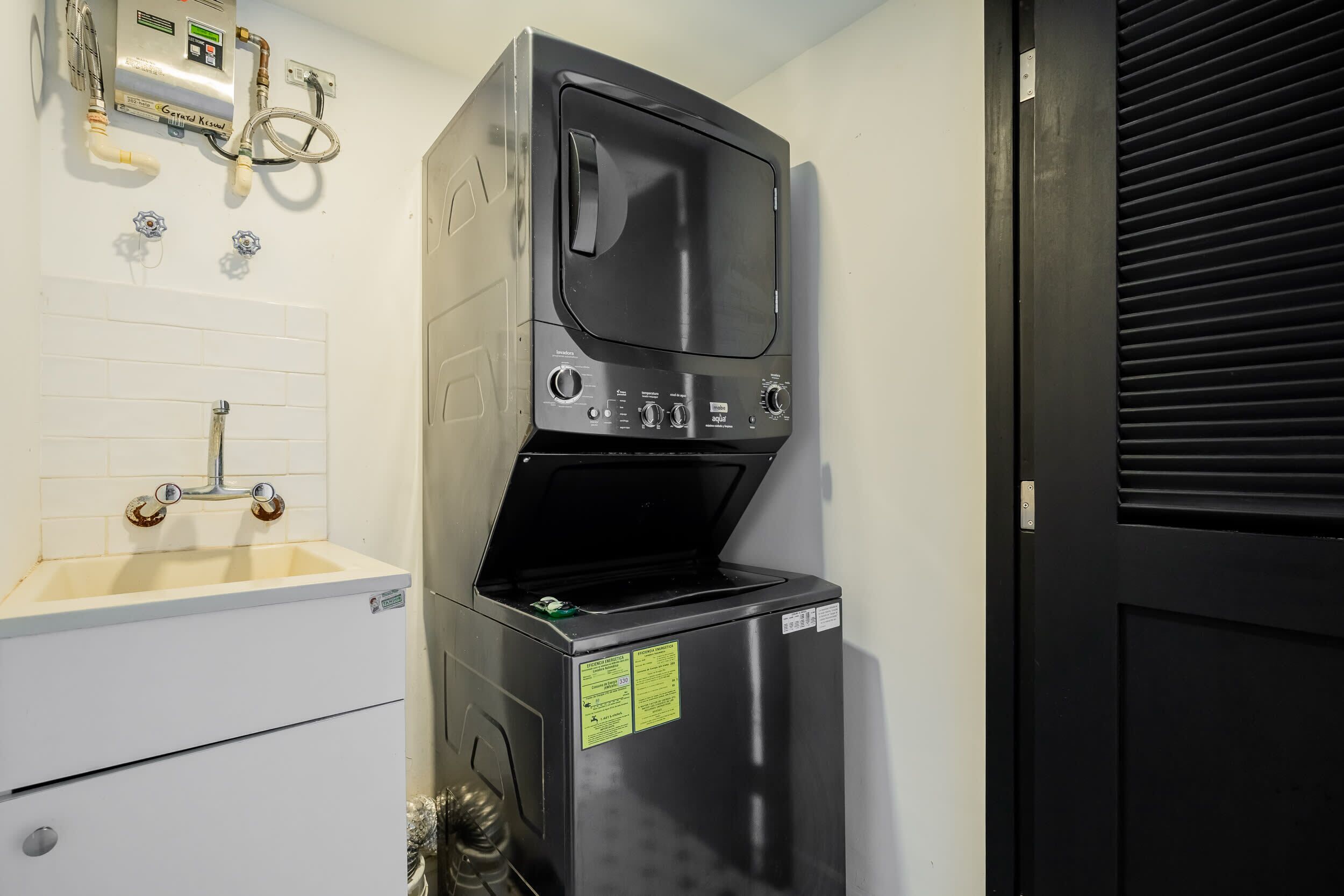 Un appareil compact combinant lave-linge et sèche-linge, en acier inoxydable, doté d’un panneau de commande, d’un petit évier avec un robinet et d’une porte avec un volet noir.