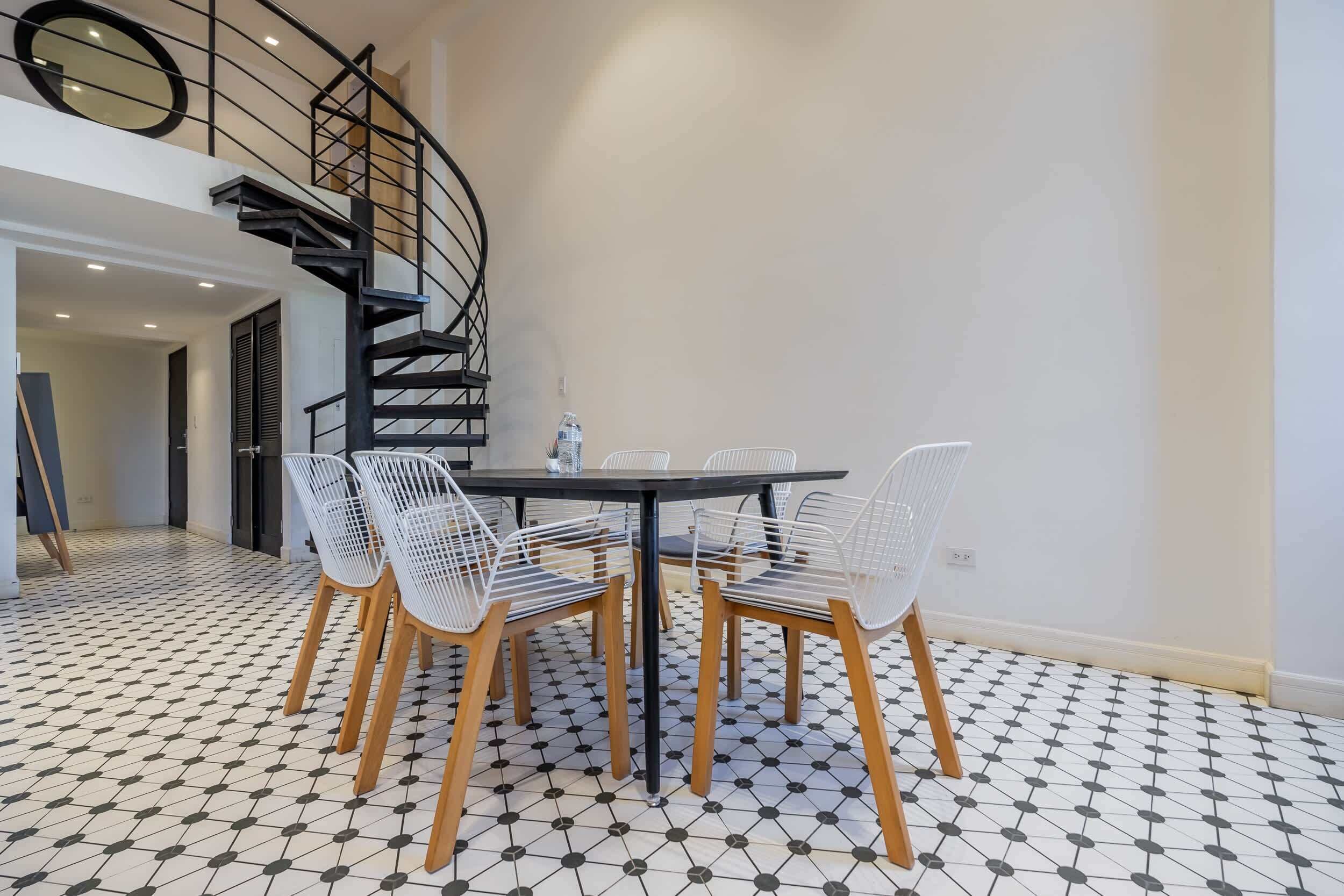 Comedor con mesa negra y sillas blancas, una escalera en espiral negra y suelo de baldosas.