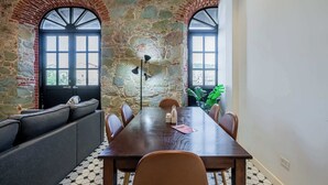 Un comedor con pared de piedra, dos ventanas arqueadas, una mesa de madera y un sofá.