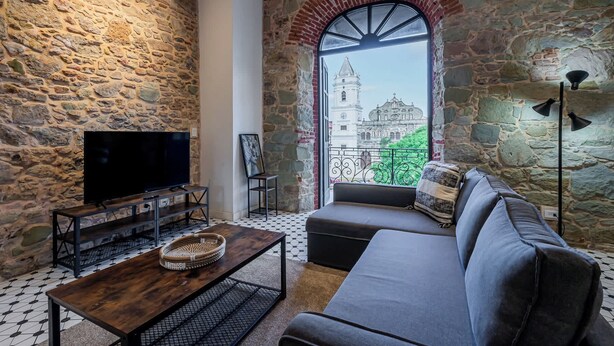 Una sala de estar con paredes de piedra, un ventanal con vistas a un edificio histórico, un televisor de pantalla plana, un sofá y una mesa de centro.