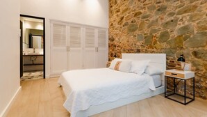 Un dormitorio con pared de piedra, una cama con ropa de cama blanca, una mesita de noche con lámpara y un armario con puertas batientes.