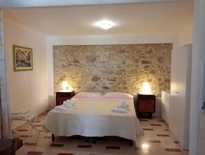 A bedroom with a stone wall, two bedside tables with lamps, a bed with a white bedspread, and a chair with a towel. - Residenza Giancesare Family Apartments (Capaccio-Paestum)