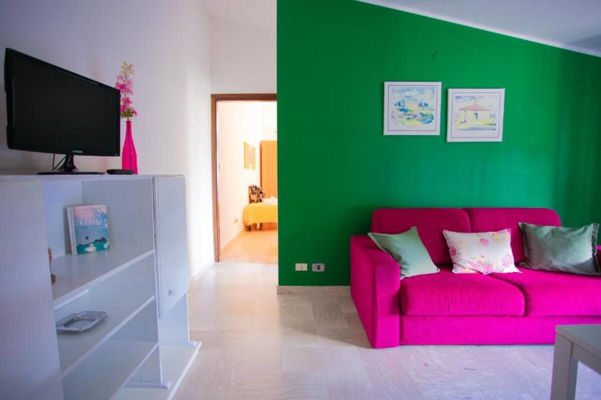Ein Zimmer mit einer leuchtend grünen Wand, einem rosafarbenen Sofa mit Kissen, einer weißen TV-Konsole und einem Bücherregal.
