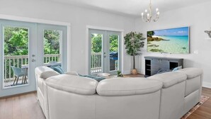 Ein geräumiges Wohnzimmer mit einem weißen Ledersofa, einem wandmontierten Flachbildfernseher und einem Balkon mit Blick auf Grünflächen.