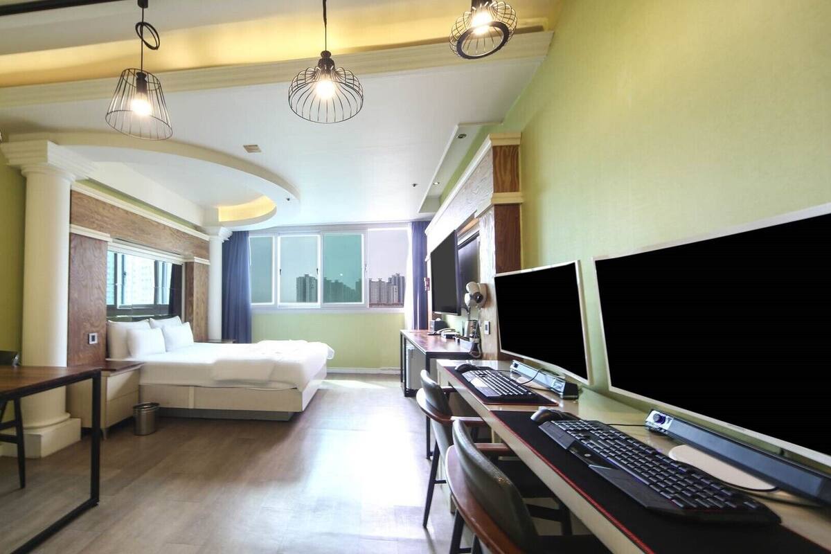 A modern hotel room with a bed, desk, computer, and large windows.
