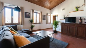 Ein Wohnzimmer mit einer Couch, einem Couchtisch und Topfpflanzen. Ein Fernseher steht auf einer Holzkommode und ein Fenster ist mit Vorhängen drapiert.