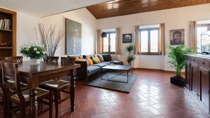 Ein geräumiger Wohnbereich mit Holzbalkendecke, einem dunkelgrauen Sofa mit Kissen, einem Esstisch mit Stühlen und einem Bücherregal.