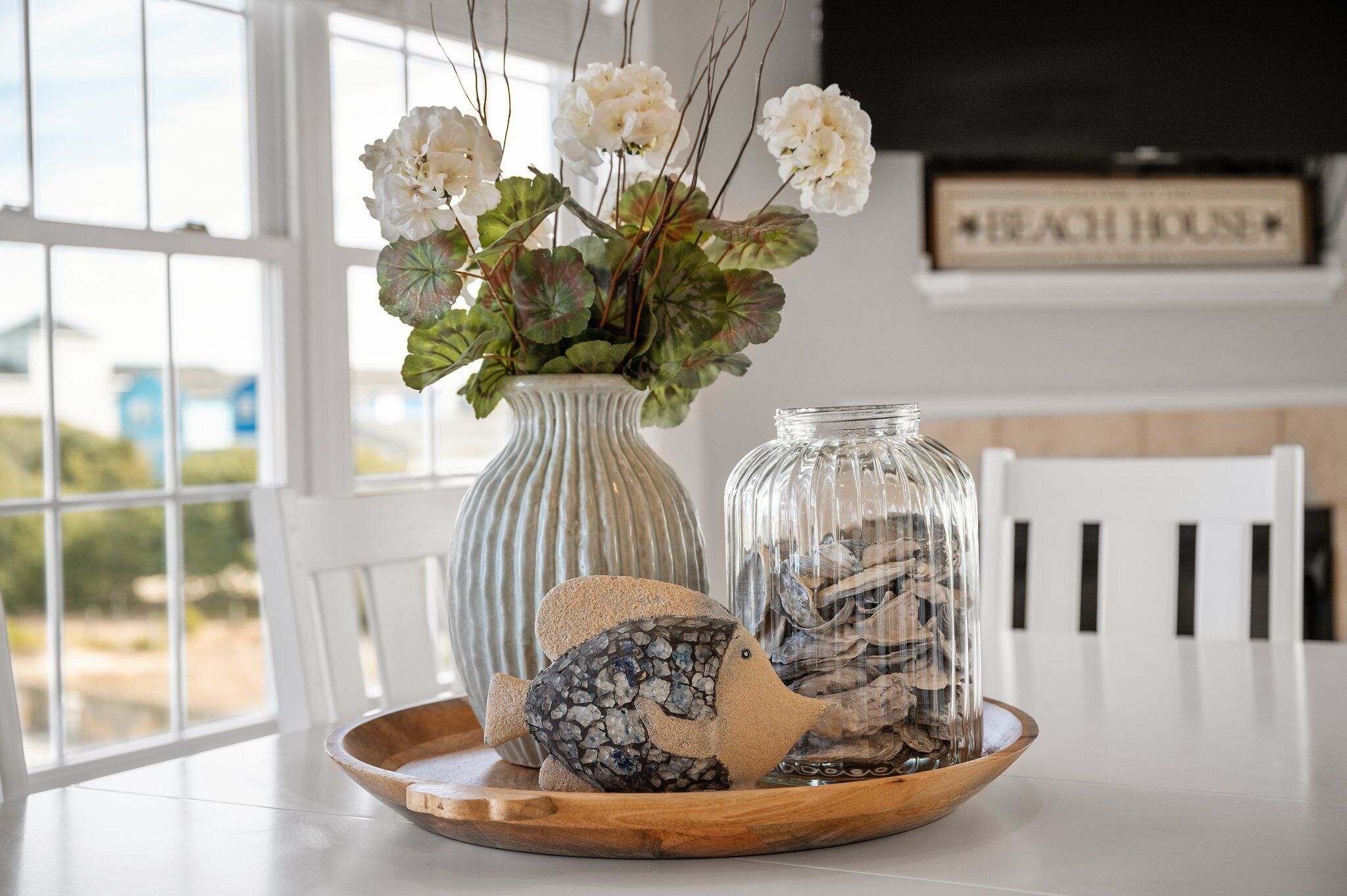 A dining table with a decorative centerpiece featuring a vase with white flowers, a glass jar with rocks, and a fish-shaped ornament.