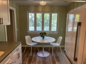 A kitchen with a round table, two white chairs, a window with a view of greenery, and a refrigerator. - Sea Palms St Simons Island, GA Decorator furnished 2/2 island escape (St. Simons Island)