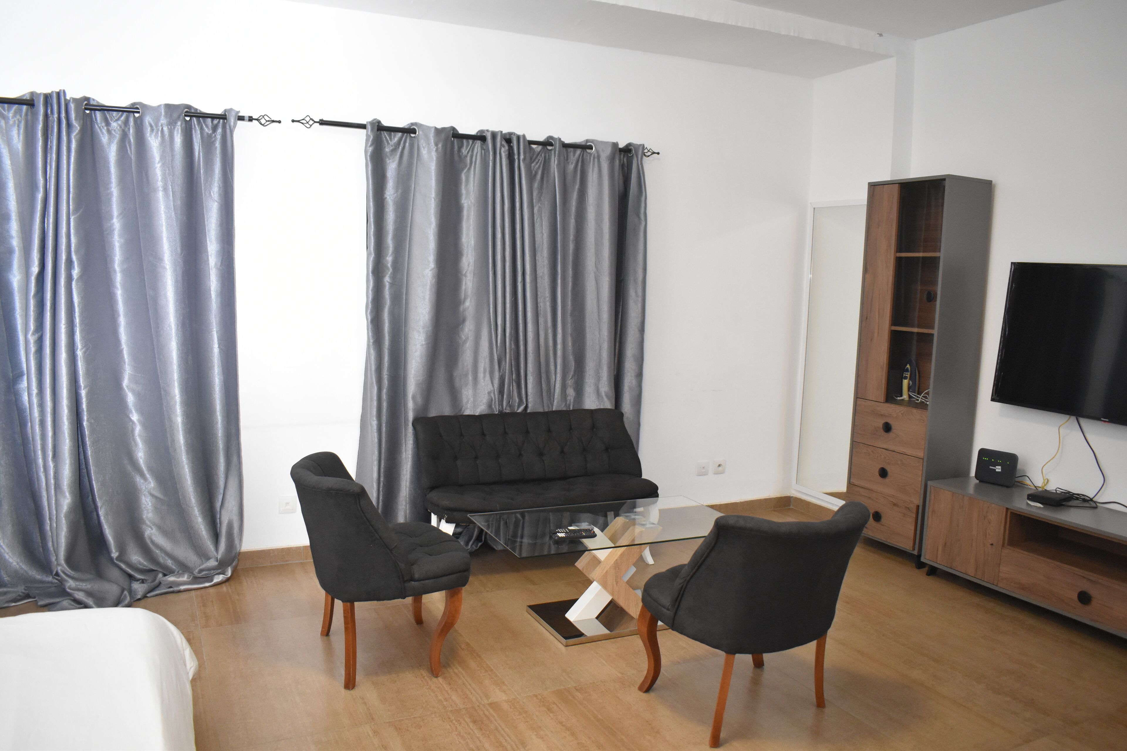 A modern living room with a black sofa, two black armchairs, a glass coffee table, and a wooden cabinet with a TV.