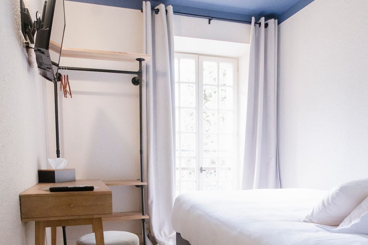 Un dormitorio con cama, escritorio, silla, ventana con cortinas y un cubo de basura.