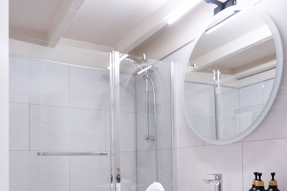 Un baño moderno con ducha envuelta en vidrio, un lavabo blanco con un espejo redondo y toallas dobladas con cuidado en un estante de madera.