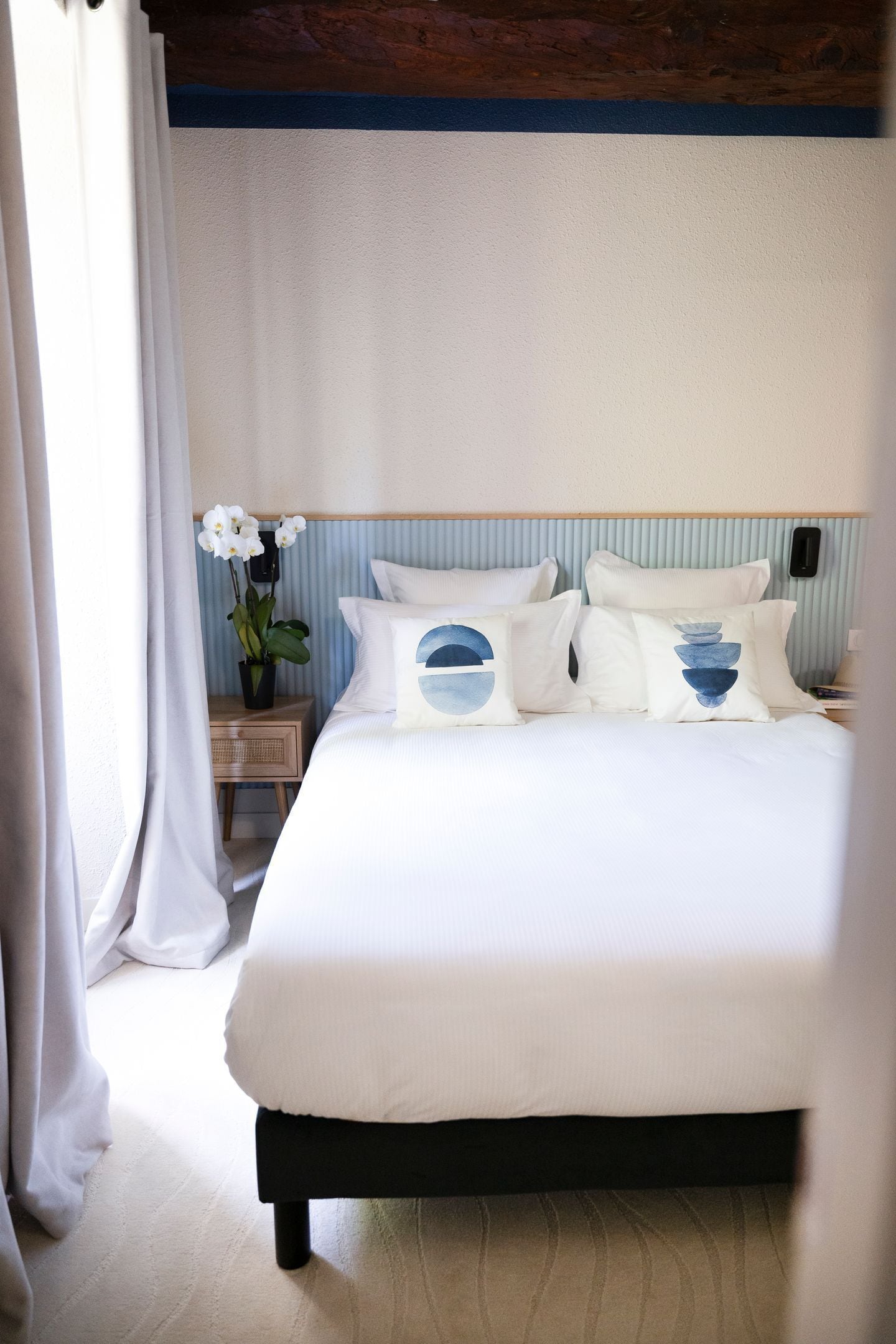 A neatly made bed with white bedding and decorative pillows, a bedside table with a vase of flowers, and a wooden headboard.