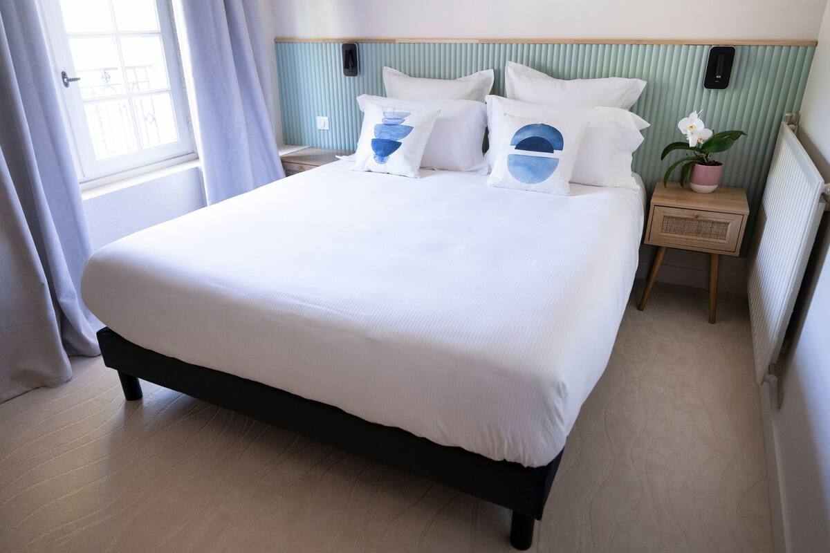 Una cama bien hecha con ropa de cama blanca y almohadillas decorativas azules, una mesita de noche con una planta pequeña en maceta y una ventana con cortinas.