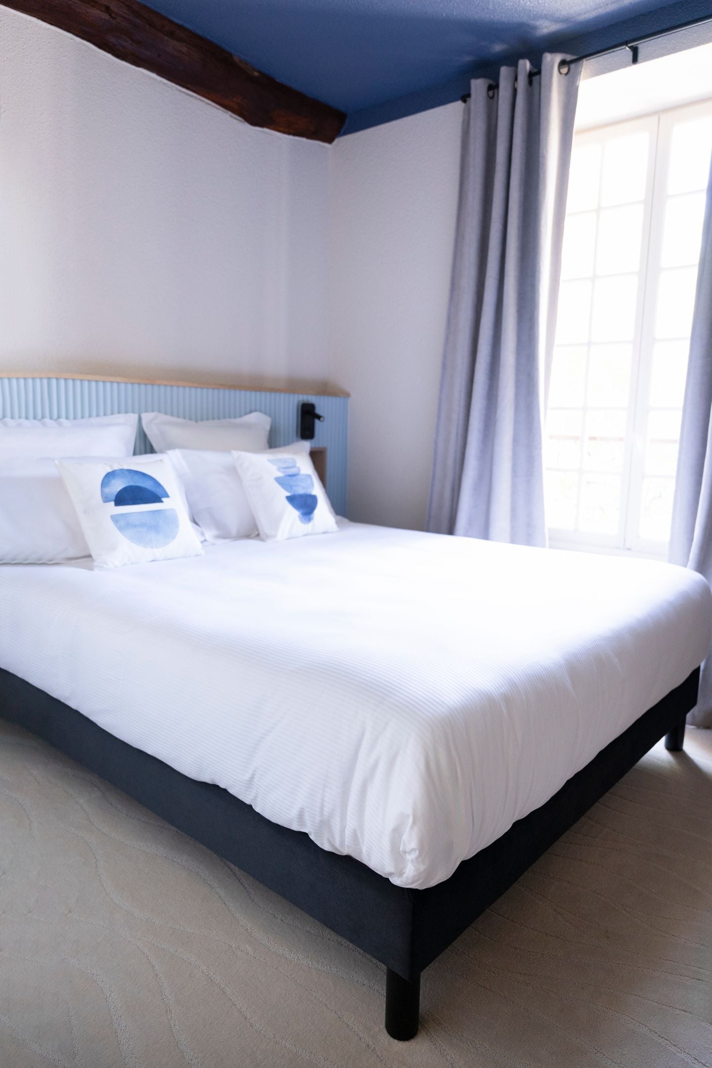 Una cama bien hecha con ropa de cama blanca y almohadas azules, un cabecero de madera y una ventana con cortinas.