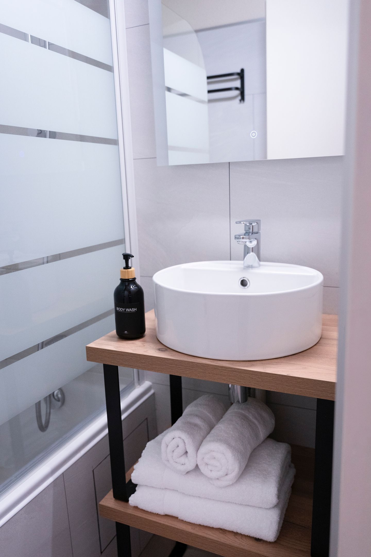 Een badkamer met een wit wastafel, een zwarte fles douchegel en twee opgerolde handdoeken op een houten plank.