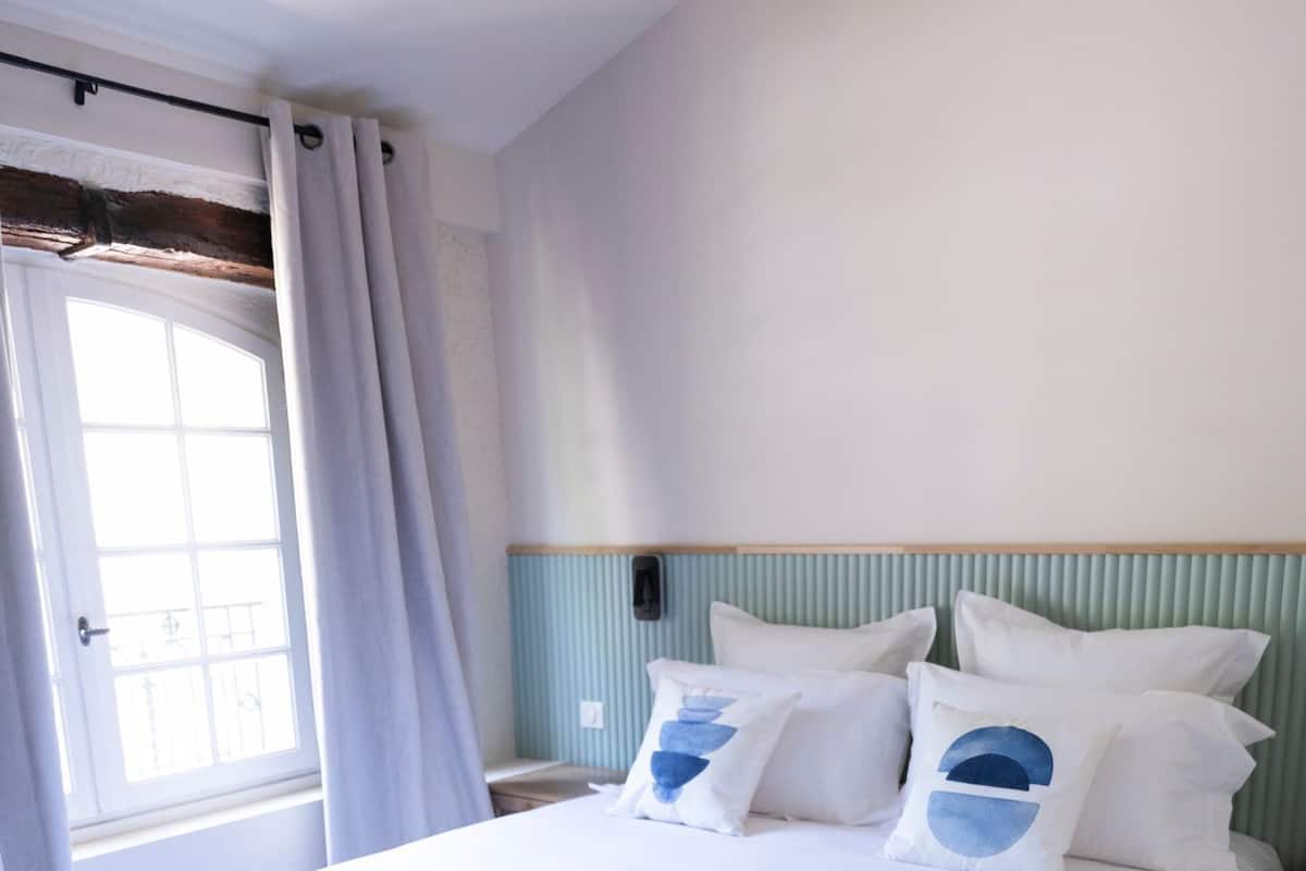 Una cama bien tendida con sábanas blancas y almohadas circulares azules, cabecera de madera, ventana con cortinas y suelo de madera.