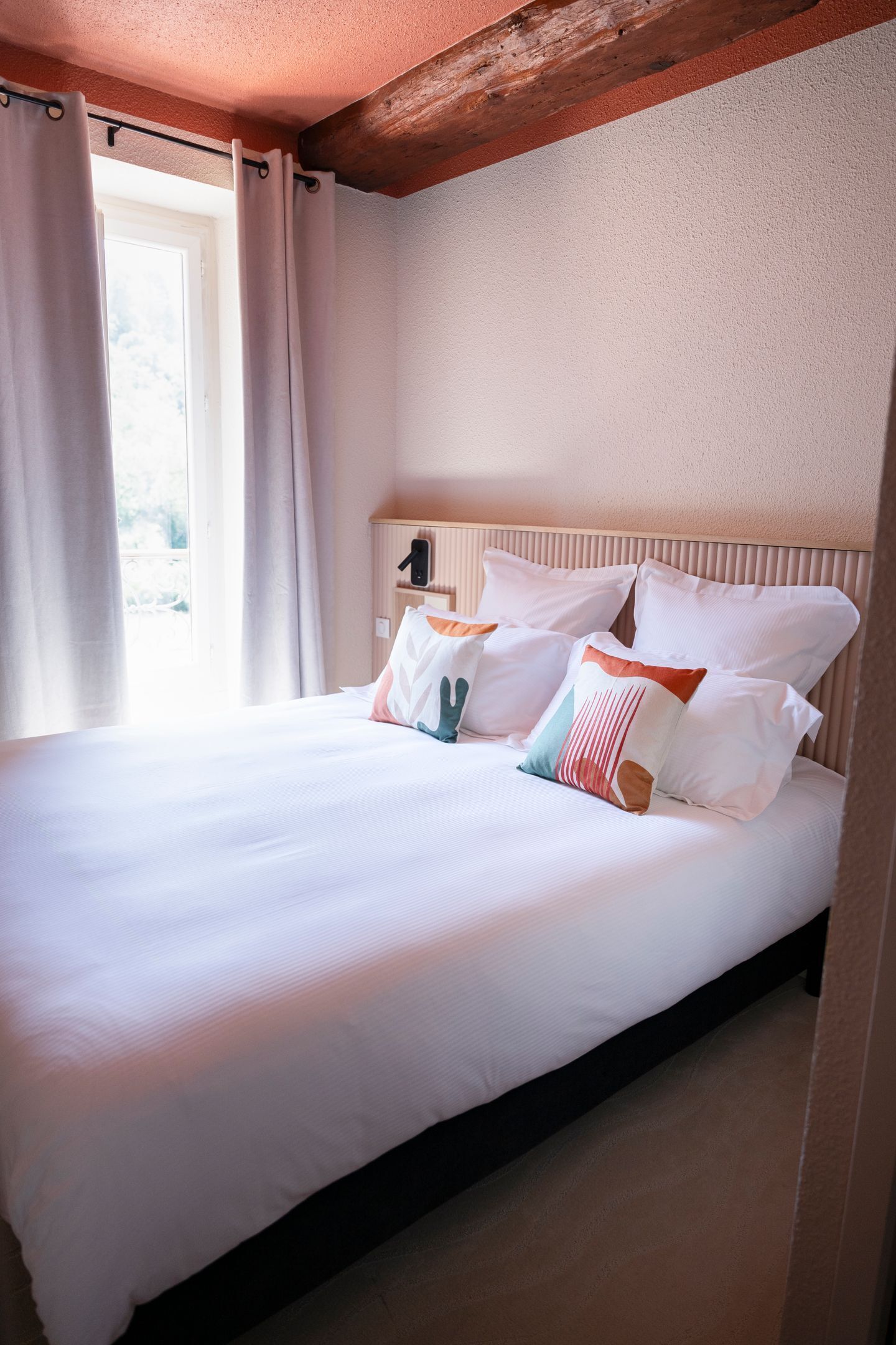 A neatly made bed with white linens and several pillows in a room with a wooden ceiling and a window with curtains.