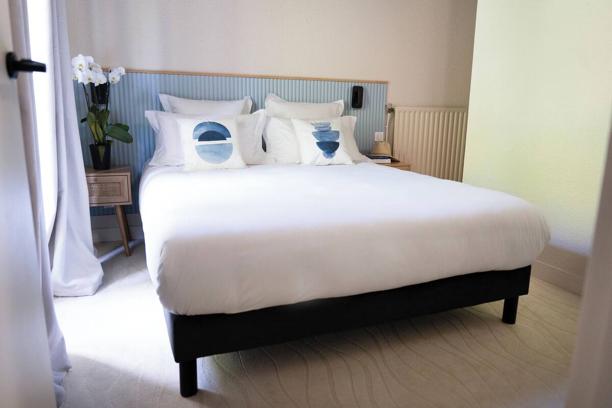 Una cama bien hecha con ropa blanca y almohadas decorativas, una mesita de noche con lámpara y una planta en maceta.