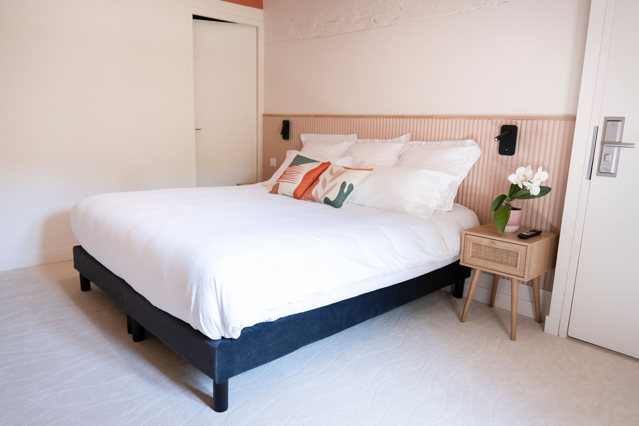 A neatly made bed with white linens and decorative pillows, a bedside table with a vase of flowers, and a wooden headboard.