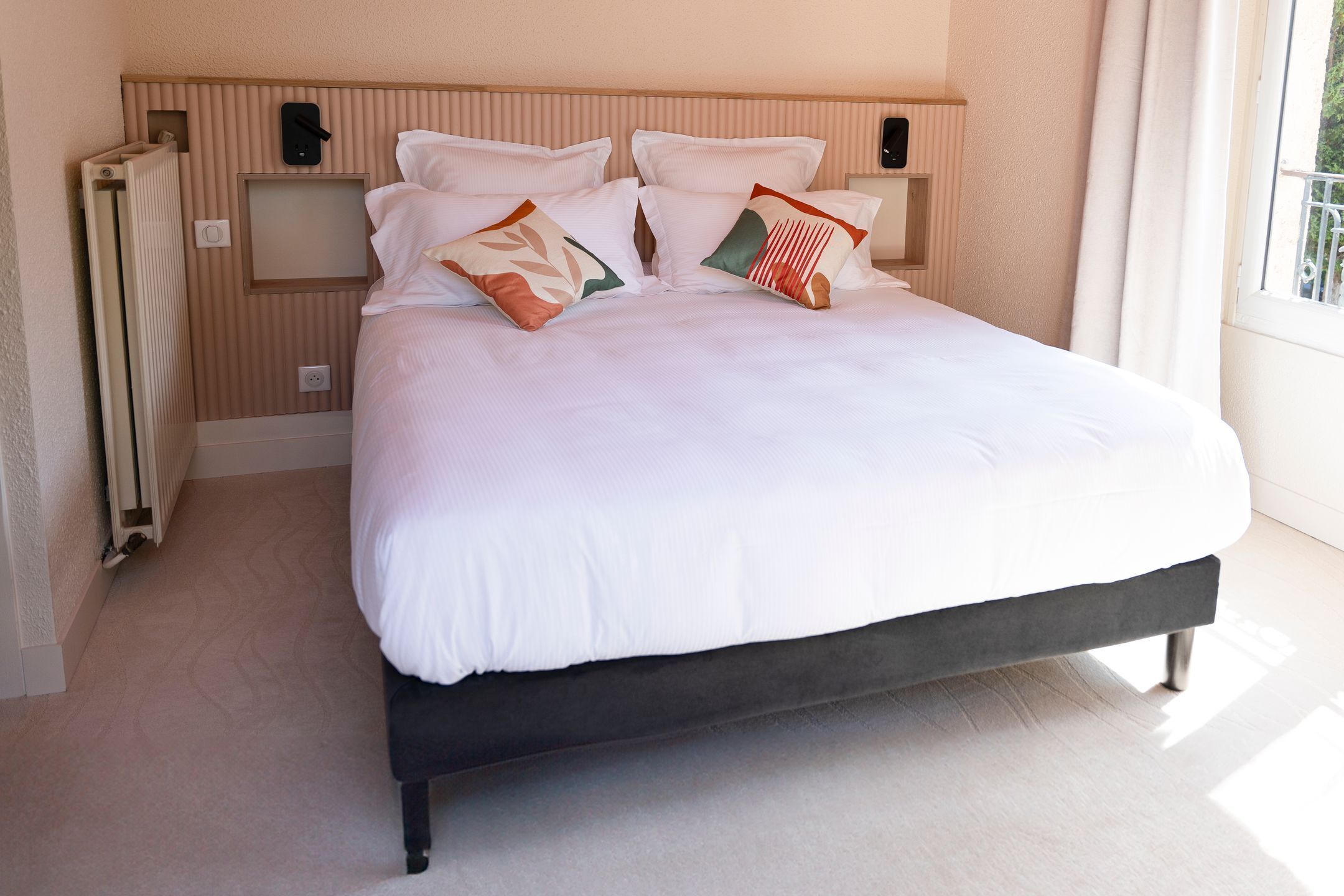 Un lit bien fait, avec une literie blanche et des coussins décoratifs, une tête de lit en bois et un radiateur sur le côté gauche.