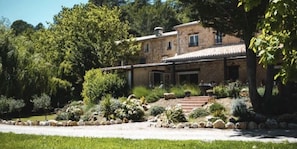 A stone house with a covered patio, surrounded by a landscaped garden with various plants and a gravel driveway.