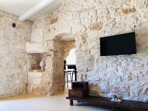 A room with exposed stone walls, a wooden bench, a small table, and a flat-screen TV mounted on the wall. - Augusta Trulli Resort (Polignano a Mare)