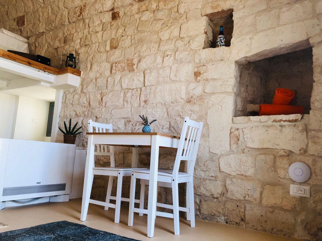 A stone-walled room with a white table and chairs, a small shelf with a lantern, and a red item in a niche.