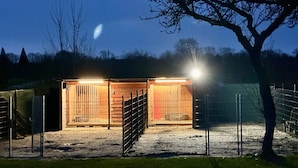 A fenced outdoor area with a wooden structure, possibly a shed or small building, illuminated by artificial lighting.