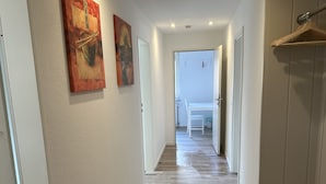 A hallway with wooden flooring, white walls, and two paintings on the left wall.