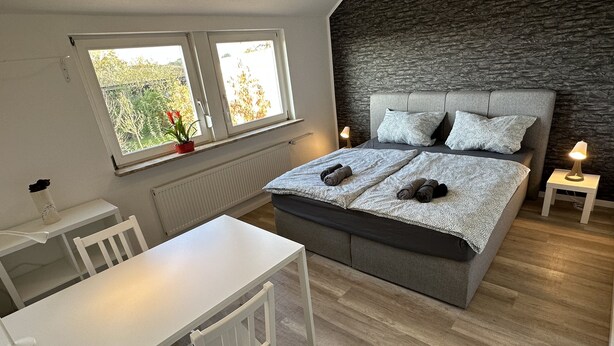 A bedroom with a bed, a desk, and a chair. There are two windows with a view of trees and a stone wall behind the bed.