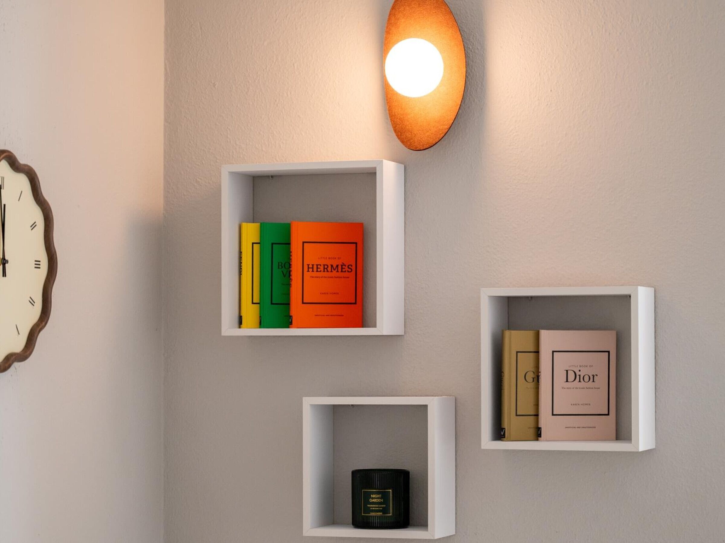 Trois étagères blanches ornées de livres et d’une bougie, une horloge murale et un luminaire fixé au mur.