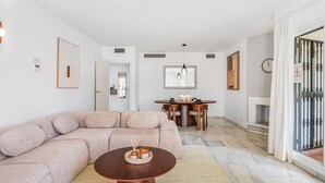Ein Wohnzimmer mit einer Eckcouch, einem runden Couchtisch, einem Essbereich mit Tisch und Stühlen sowie einer durchgehenden Tür erkennbaren Küche.
