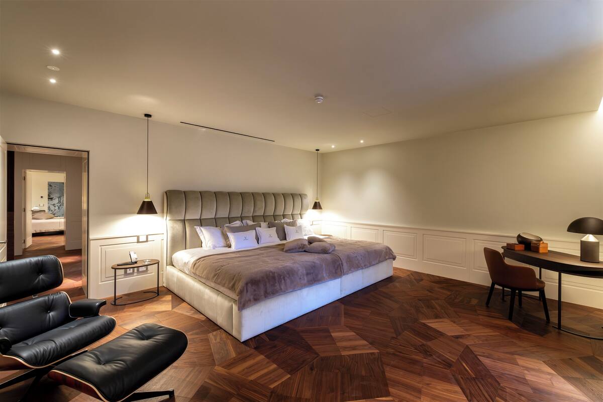 A modern bedroom with a large bed, a sitting area with a chair, a small table, and a desk with a lamp.