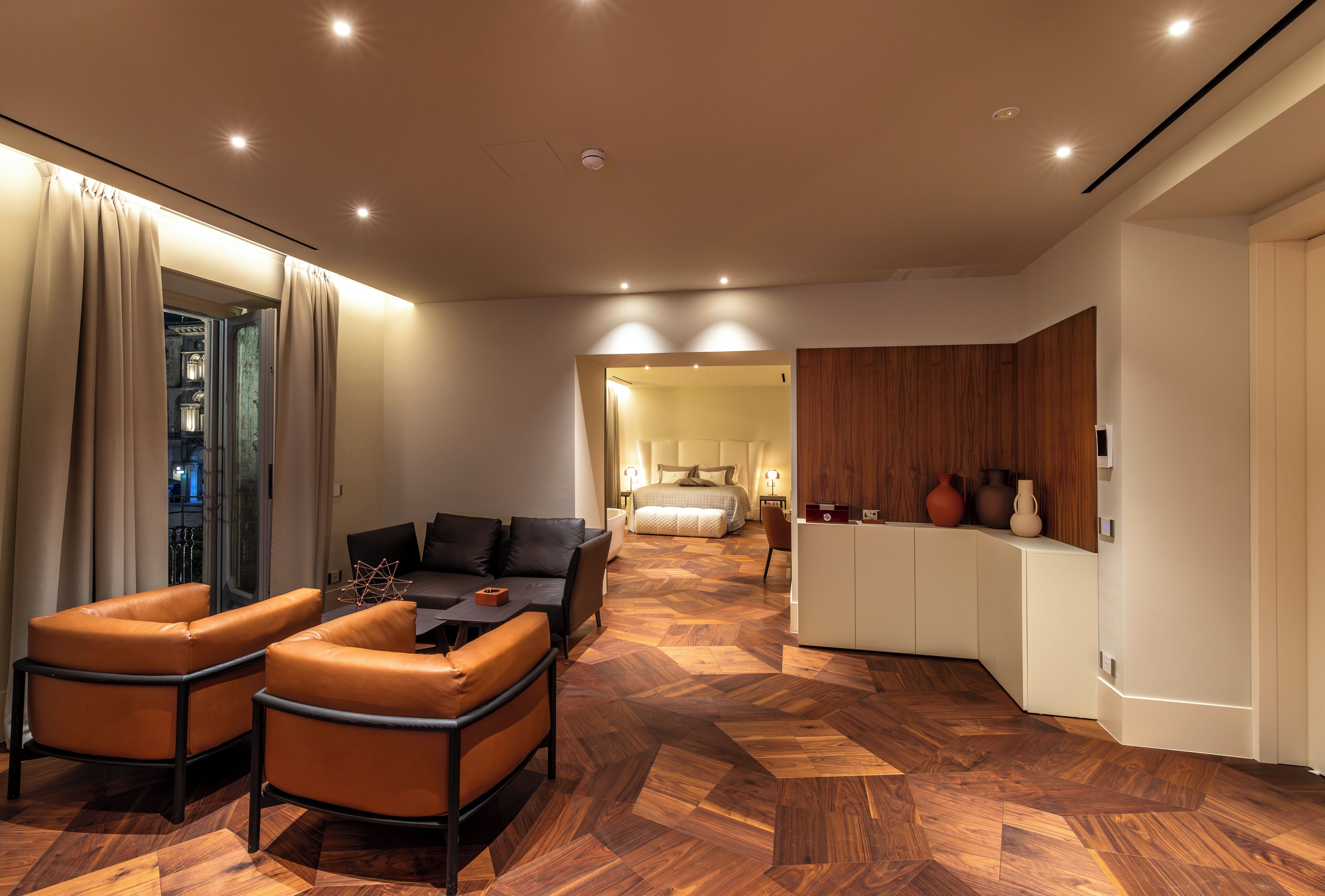 A modern hotel room with a wooden herringbone floor, a sofa, a dining area, and a bedroom with a bed and nightstands.