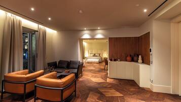 A modern hotel room with a wooden herringbone floor, a sofa, a dining area, and a bedroom with a bed and nightstands.