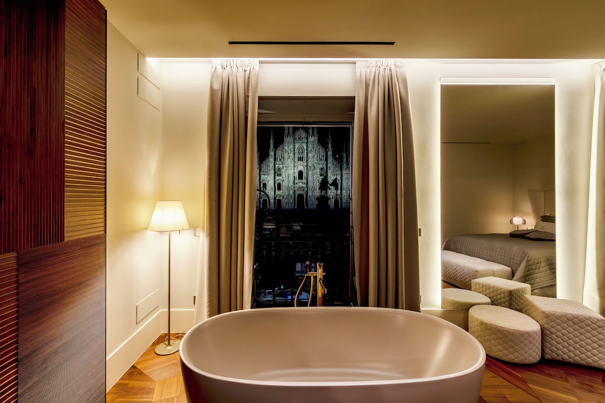 A modern bathroom with a large bathtub, a wooden panel wall, and a view of a cathedral through a window.