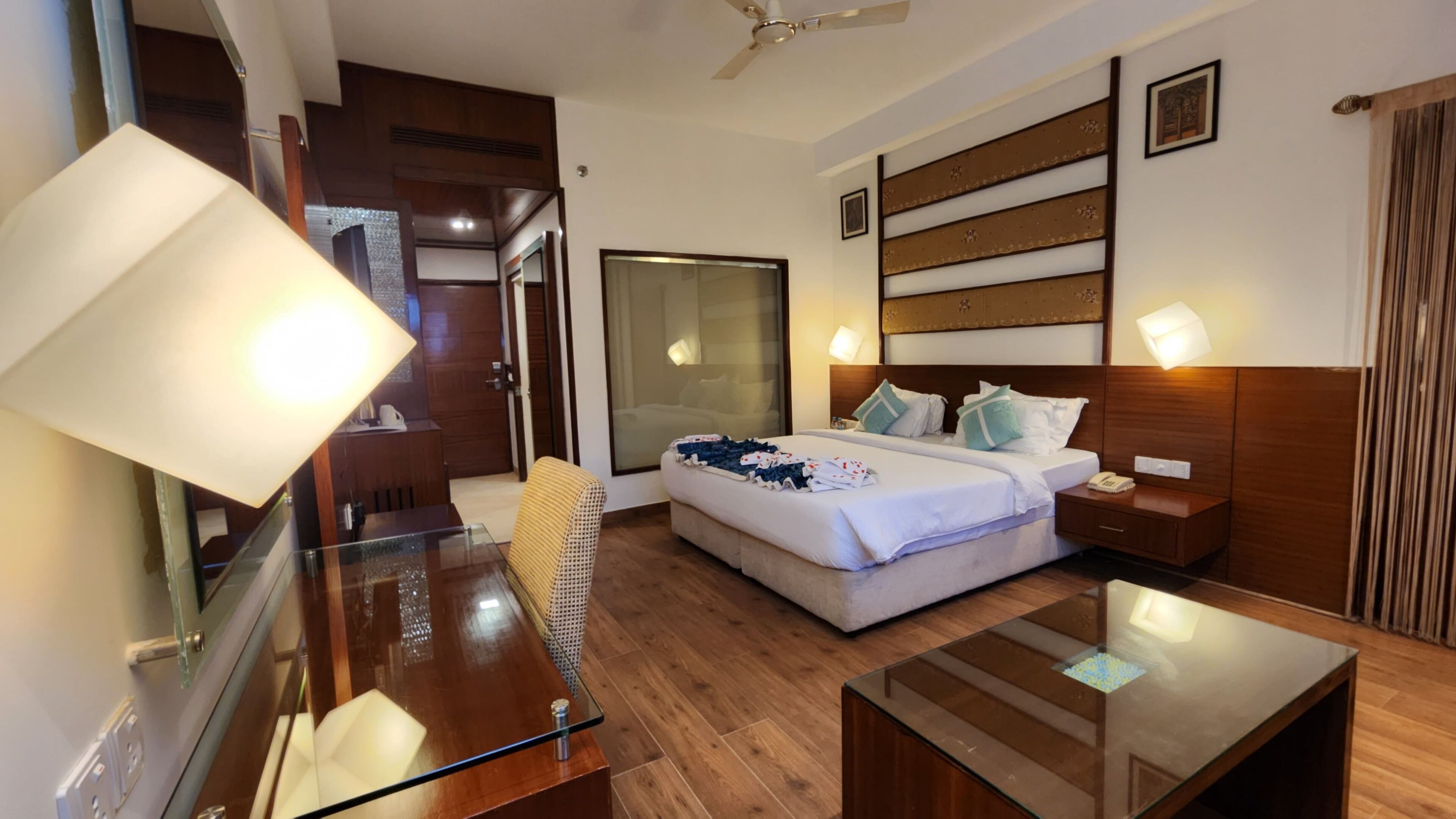 Habitación de hotel con cama, mesitas de noche, una mesa de centro de vidrio, un espejo, ventilador de techo y una puerta corredera.
