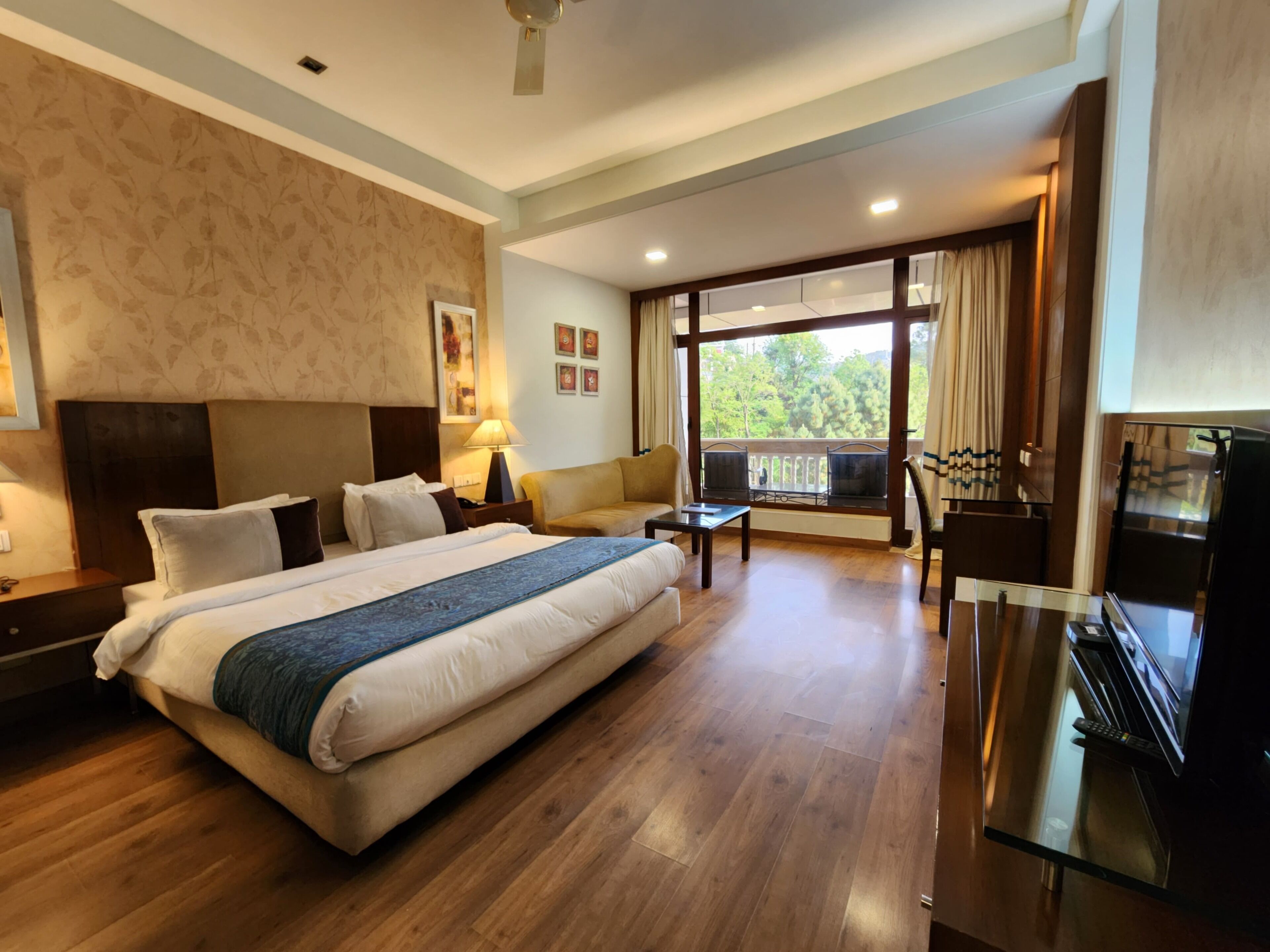 A spacious hotel room with a large bed, a sofa, a TV, and a balcony with a view of the outdoors.