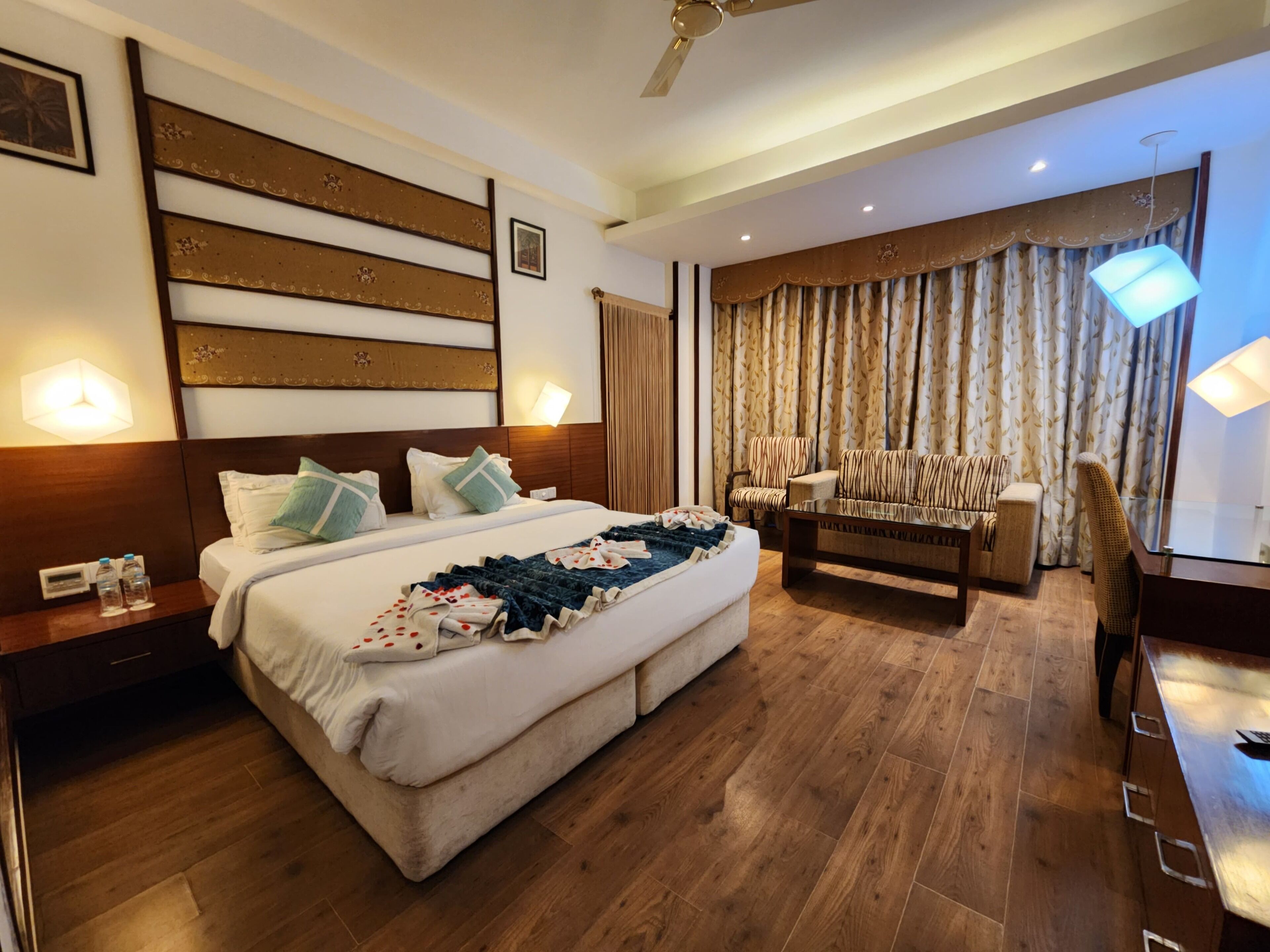 Une chambre d’hôtel avec un grand lit, un canapé, un bureau et une chaise. Il y a un ventilateur de plafond et une fenêtre avec des rideaux.
