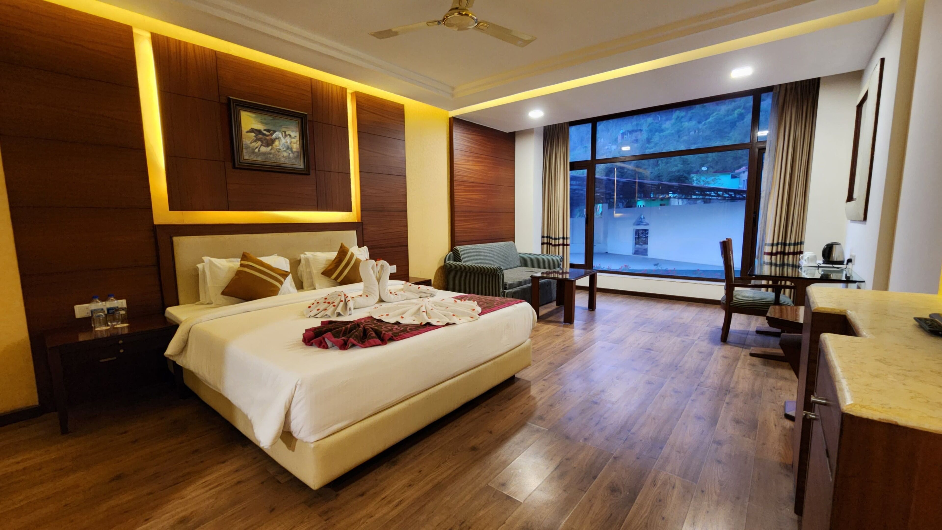 A hotel room with a large bed, a sofa, a dining table, and a view of the cityscape through large windows.