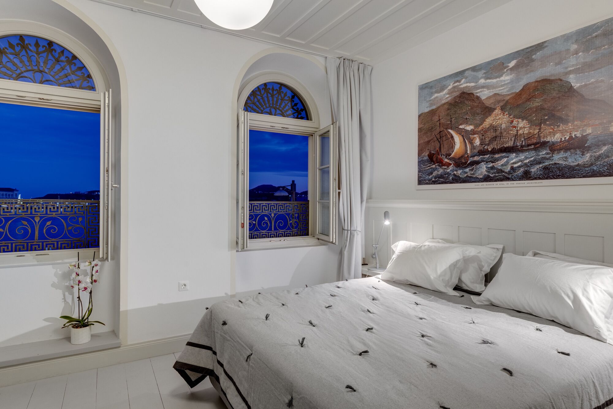A bedroom with a large bed, two windows with decorative grills, a painting of a ship on the wall, and a potted orchid.