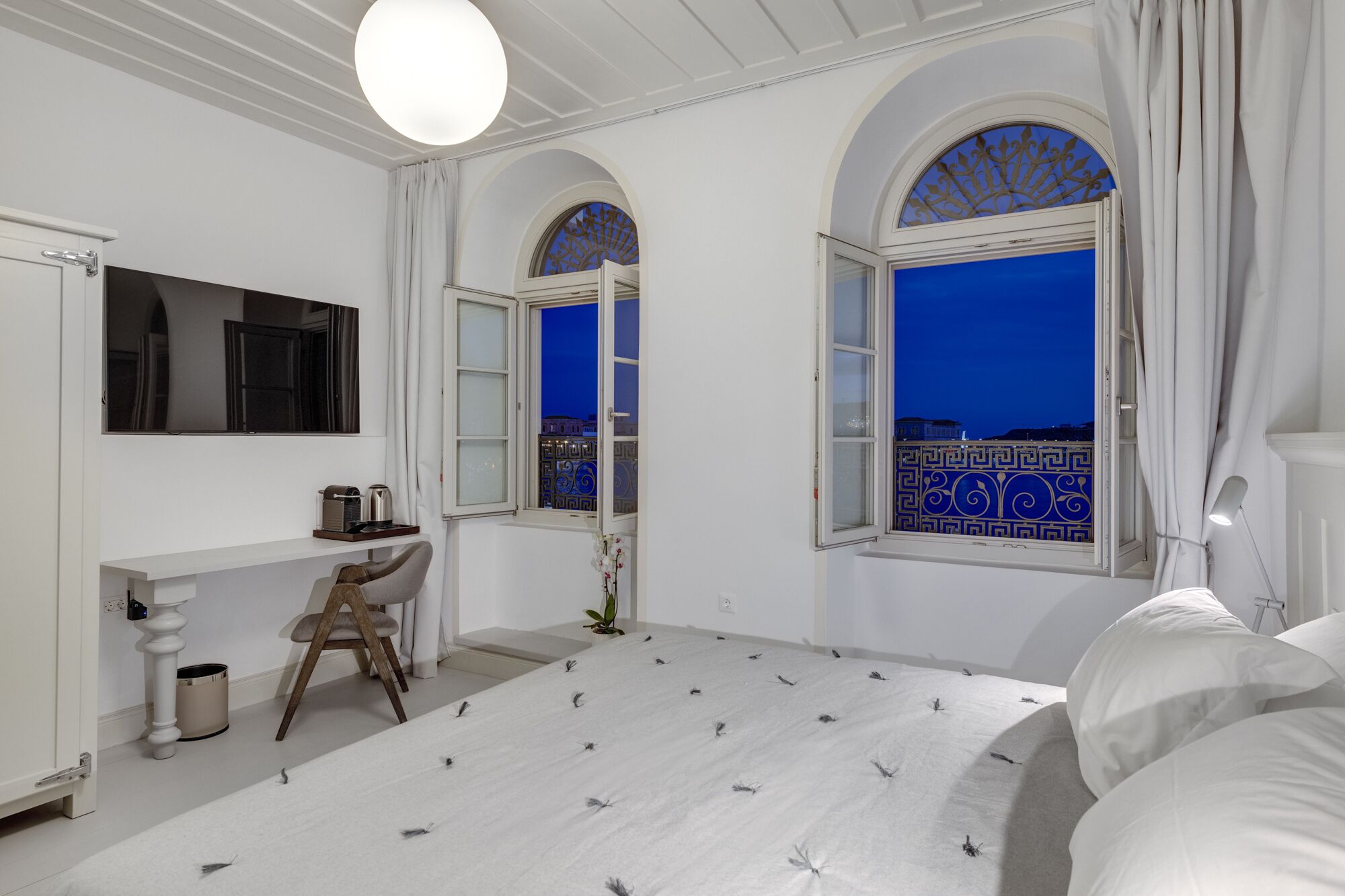 Un dormitorio con cama, escritorio, silla y dos ventanas con detalles decorativos.