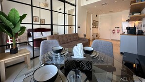 A modern living room with a glass dining table set for two, a sofa, and a large green plant.