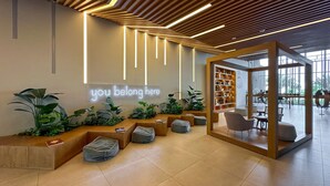 A modern reception area with a welcoming sign, wooden seating, a plant-filled corner, and a view into the adjacent room.