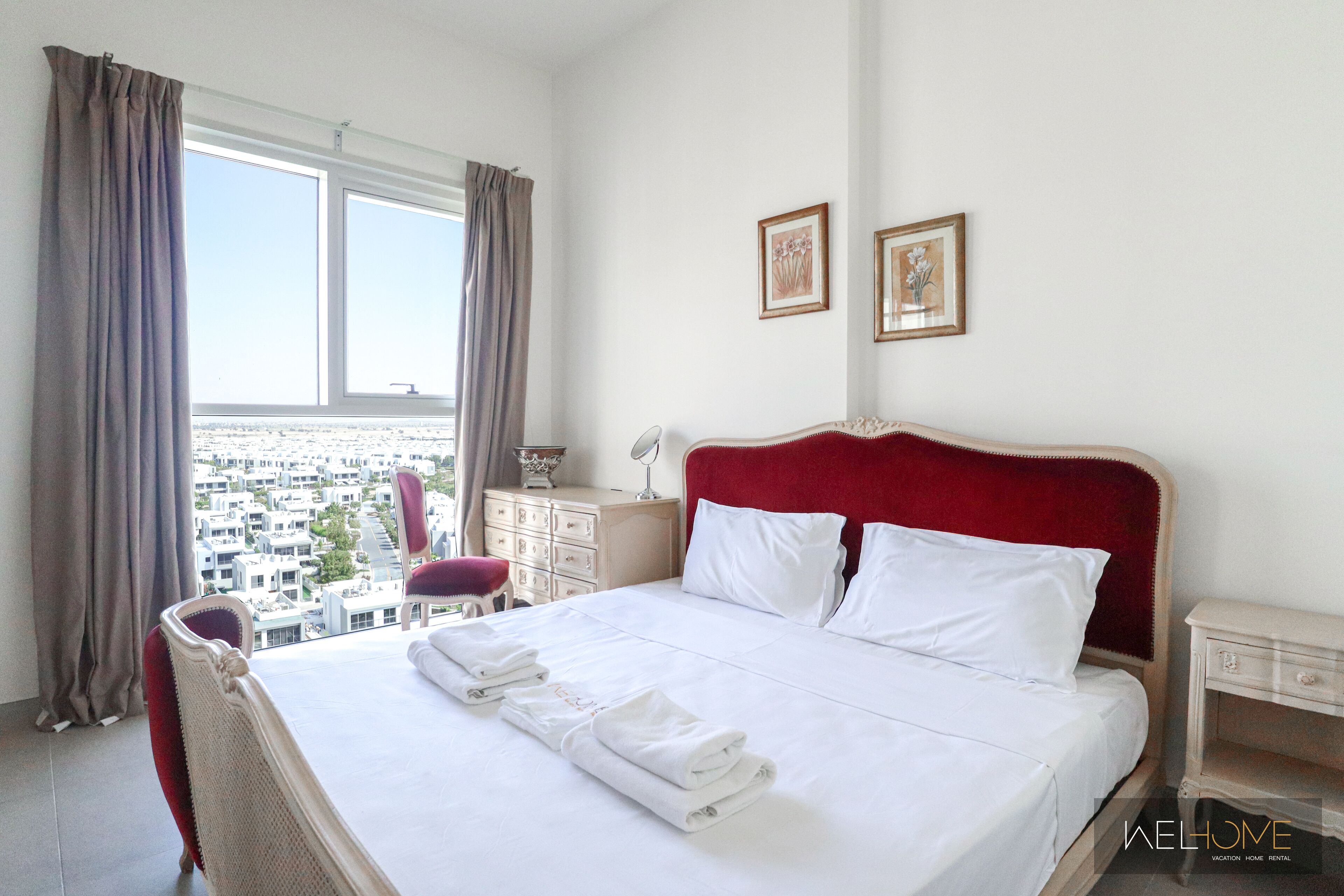 A hotel room with a large bed, a red headboard, white bedding, a view of the city, a small table with a lamp, and two framed pictures on the wall.