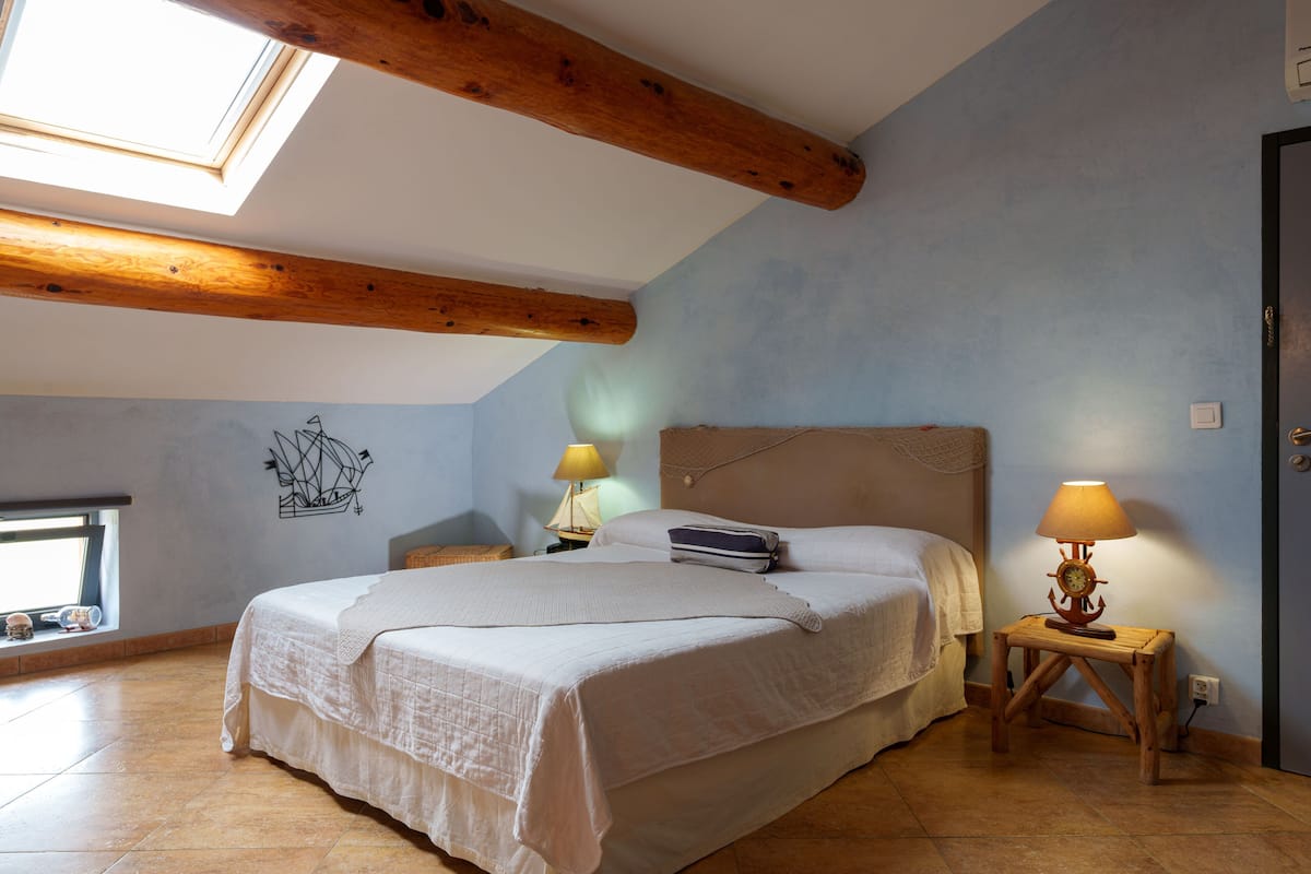 A bedroom with a large bed, wooden beams, a skylight, a wall-mounted ship decoration, two bedside tables with lamps, and a window with a view.